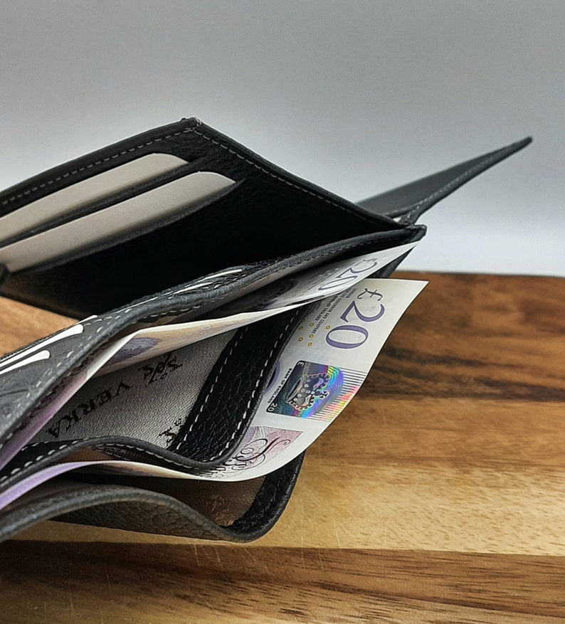 Puede incluir: Una cartera de cuero negro con detalles de costura gris est&aacute; abierta y llena de billetes de veinte libras.