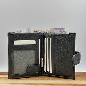 Puede incluir: Una cartera de cuero negro con m&uacute;ltiples ranuras para tarjetas y un compartimento con cremallera. La cartera est&aacute; abierta y tiene un billete de veinte libras dentro.