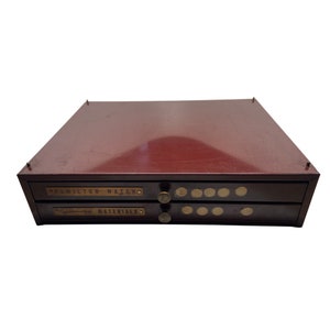 May include: A two-drawer metal storage box with a maroon finish. The top drawer has a brass label that reads "Hamilton Watch" and the bottom drawer has a brass label that reads "Gem Materials".