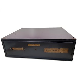 May include: A black metal drawer unit with a single drawer. The drawer has a brass label that reads "SYSTEM 100" and a paper label that reads "BESTFIT System # 300".