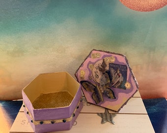 以下が含まれることがあります： 金色に輝く内側と装飾的な蓋が付いた、紫色の六角形の箱。蓋には、金色のアクセントが施された蝶と、ピンク色の背景に「Spread Your Wings」という文字が書かれています。