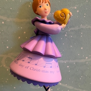 May include: A decorative Christmas ornament featuring a figurine of a woman in a purple dress holding a gold heart with the number 9. The dress has musical notes and the text "9th day of Christ-mas my true".