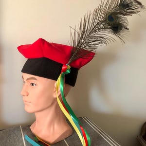 May include: A red felt hat with a black brim and a peacock feather. The hat has four long ribbons attached to it in the colors red, yellow, green, and blue.