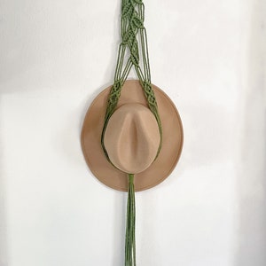 May include: A green macrame hat hanger with a wooden ring at the top and a long tassel. A brown hat is hanging on the hanger.