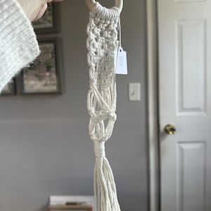 May include: A white macrame plant hanger with a wooden ring at the top and a glass test tube in the center. The hanger has a tassel at the bottom. The tag attached to the hanger says "HGH".