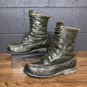 May include: A pair of dark green leather work boots with laces and metal eyelets. The boots have a moccasin-style toe and a thick, lugged sole. The boots are worn and show signs of use.