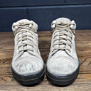 May include: A pair of high-top sneakers in a light gray suede material with matching gray laces and a black rubber sole. The tongue of each shoe features a circular patch with the letter A in the center. The design includes a rounded toe cap.