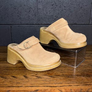 May include: Tan suede clogs with a rounded toe and a thick wooden heel. The clogs have a strap across the top with a metal buckle. The shoes are displayed on a clear plastic box on a wooden surface.