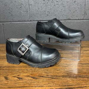 May include: A pair of black leather ankle boots with a chunky sole. One boot features a silver buckle with the brand name. The boots are displayed on a wooden surface with a gray brick background.