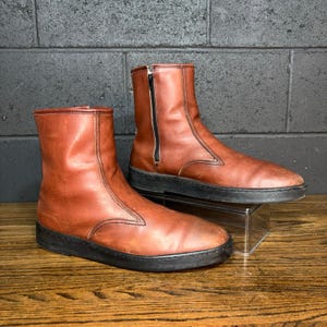 May include: A pair of brown leather ankle boots with black soles. The boots have a zipper on the side and are displayed on a clear acrylic stand. The background is a dark brick wall and a wooden surface.