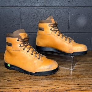 May include: A pair of tan leather hiking boots with brown accents and black soles. The boots have brown laces and the brand name "AVOLO" is visible on the side. The boots are displayed on a wooden surface.