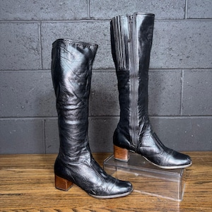 May include: A pair of black leather knee-high boots with wooden block heels. One boot has a zipper running up the side. The boots are displayed on a wooden surface against a dark brick wall.