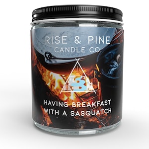 Könnte beinhalten: Ein Glas-Kerzenglas mit schwarzem Deckel. Das Etikett trägt den Text "RISE & PINE CANDLE CO." und "HAVING BREAKFAST WITH A SASQUATCH" über einer Lagerfeuerszene. Die Kerze hat einen weißen Hintergrund.