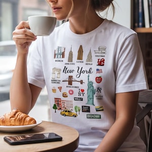 A woman wearing a white t-shirt with New York City landmarks printed on it is sitting at a table in a cafe, sipping coffee and enjoying a croissant.
