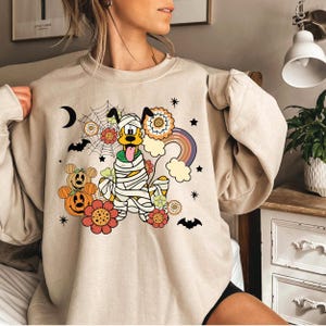 May include: A beige sweatshirt with a cartoon image of a yellow dog dressed as a mummy. The dog is surrounded by flowers, a rainbow, stars, and bats. The dog has a tongue sticking out and is wearing a white bandage around its head. The dog is surrounded by a spiderweb and a crescent moon. The sweatshirt has a relaxed fit and long sleeves.