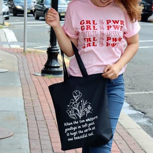 May include: A black tote bag with a white floral design and the text 'When the sun whispers goodbye in soft gold, it hugs the earth as one beautiful soul.' The bag is being held by a person wearing a pink t-shirt with the text 'GRL PWR' repeated in red letters.