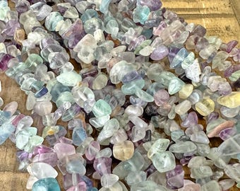 May include: A close-up of a collection of tumbled fluorite gemstones. The stones are a mix of colors, including purple, green, blue, and clear. The gemstones are irregular in shape and size, and appear to be strung together.