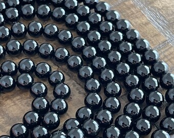 Puede incluir: Una hebra de cuentas de ónix negro, cada cuenta es lisa y pulida, creando un aspecto uniforme. Las cuentas están dispuestas en un patrón circular, mostrando su acabado brillante.