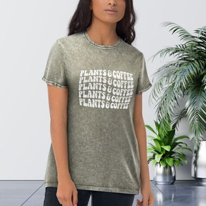 Op de afbeelding: Een groen, distressed, t-shirt met korte mouwen met de tekst "PLANTS & COFFEE" die in een retro-lettertype wordt herhaald.