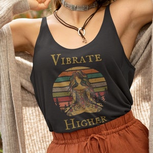 Pode incluir: Um top preto de alças com um gráfico de estilo vintage de uma mulher meditando com o texto "Vibrate Higher".