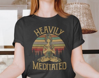 May include: A dark gray t-shirt with a vintage-style graphic of a woman meditating in a lotus pose. The graphic features a sunset background with the text "Heavily Meditated" in yellow.