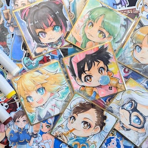 Może przedstawiać: Kolekcja kwadratowych wydruków artystycznych w stylu anime, przedstawiających różne postacie z dużymi oczami i kolorowymi włosami. Wydruki otoczone są złotą obwódką i ułożone na powierzchni z innymi ilustracjami i materiałami artystycznymi.