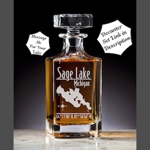 May include: A clear glass decanter with a square base and a stopper. The decanter is engraved with the words "Sage Lake Michigan" and the coordinates "44°21'06" N, 83°56'46" W".