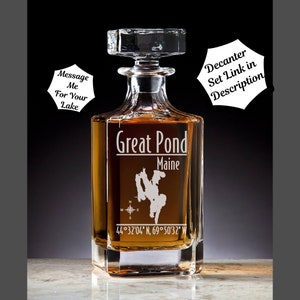 May include: A clear glass decanter with a square base and a stopper. The decanter is etched with the words "Great Pond Maine" and an outline of a lake. The coordinates "44°32′04″ N, 69°50'32" W" are also etched on the decanter.