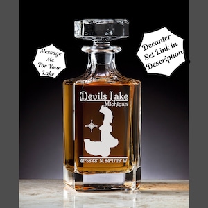 May include: A clear glass decanter with a square base and a stopper. The decanter is etched with the words "Devils Lake Michigan" and the coordinates "41°58′48″ N, 84°17'19" W".