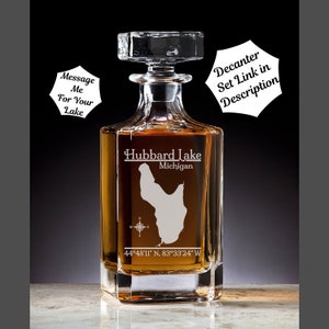 May include: A clear glass decanter with a square base and a stopper. The decanter is engraved with the outline of Hubbard Lake, Michigan, and the coordinates 44°48′11″ N, 83°33'24" W. The text "Hubbard Lake Michigan" is also engraved on the decanter.