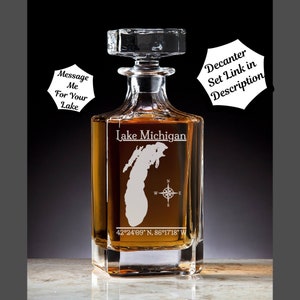 May include: A clear glass decanter with a square base and a stopper. The decanter is etched with the outline of Lake Michigan, a compass, and the coordinates 42°24'09" N, 86°17'18" W. The text "Lake Michigan" is also etched on the decanter.