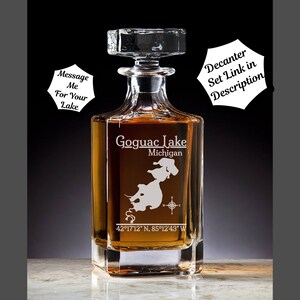 May include: A clear glass decanter with a square base and a stopper. The decanter is engraved with the words "Goguac Lake, Michigan" and the coordinates "42°17′12″ N, 85°12'43" W".