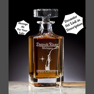 May include: A clear glass decanter with a square base and a stopper. The decanter is etched with an outline of the Detroit River in Michigan and the coordinates 42°10′19″ N, 83°07'39" W. The text "Detroit River" and "Michigan" are also etched on the decanter.