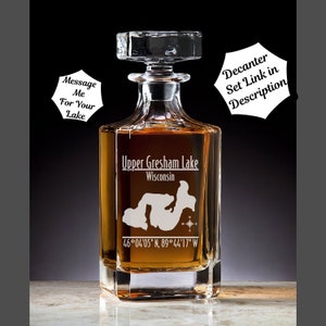 May include: A clear glass decanter with a square base and a stopper. The decanter is engraved with the words "Upper Gresham Lake, Wisconsin" and the coordinates "46°04'05" N, 89°44'17" W".
