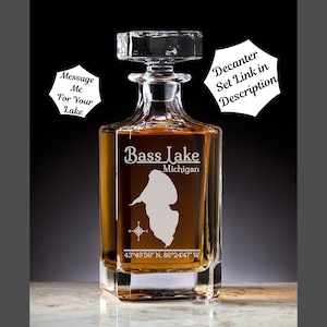 May include: A clear glass decanter with a square base and a stopper. The decanter is etched with the words "Bass Lake Michigan" and the coordinates "43°49′50″ N, 86°24'47" W".