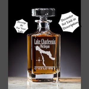 May include: A clear glass decanter with a square base and a stopper. The decanter is etched with the outline of Lake Charlevoix, Michigan, and the coordinates 45°16′26″ N, 85°09'06" W. The text "Lake Charlevoix, Michigan" is also etched on the decanter.