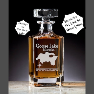 May include: A clear glass decanter with a square base and a stopper. The decanter is engraved with the words "Goose Lake, Michigan" and the coordinates "42°03′30″ N, 84°20'28" W".