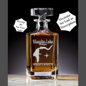 May include: A clear glass decanter with a square base and a stopper. The decanter is engraved with the words "Hamlin Lake, Michigan" and the coordinates "44°02′44″ N, 86°28'17" W".