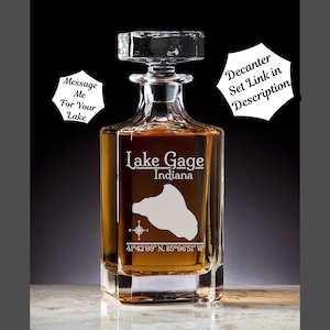 May include: A clear glass decanter with a square base and a stopper. The decanter is engraved with the text "Lake Gage Indiana" and the coordinates "41°42′09″ N, 85°06'51" W".