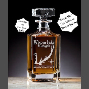May include: A clear glass decanter with a square base and a stopper. The decanter is engraved with an outline of Wixom Lake, Michigan, and the coordinates 43°49′04″ N, 84°22'38" W. The text "Message Me For Your Lake" is also engraved on the decanter.