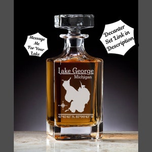 May include: A clear glass decanter with a square base and a stopper. The decanter is etched with an outline of Lake George, Michigan, and the coordinates 41°45'45" N, 85°00'43" W. The text "Lake George" and "Michigan" are also etched on the decanter.