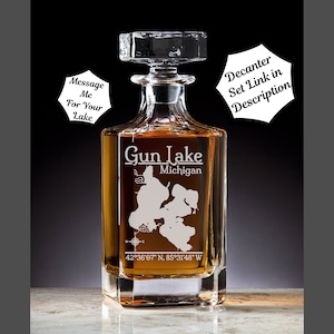 May include: A clear glass decanter with a square base and a stopper. The decanter is engraved with the words "Gun Lake, Michigan" and the coordinates "42°36′07″ N, 85°31'48" W".