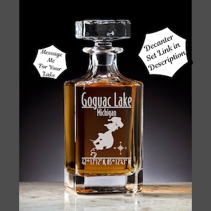 May include: A clear glass decanter with a square base and a stopper. The decanter is engraved with the words "Goquac Lake, Michigan" and the coordinates "42°17'12" N, 85°12'43" W".