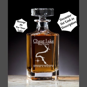May include: A clear glass decanter filled with amber liquid, featuring the text "Cheat Lake West Virginia" and a lake outline. Coordinates 39°39'48" N, 79°51'25" W are displayed below the lake graphic. The decanter has a square stopper.