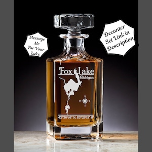 May include: A clear glass decanter with a square base and a stopper. The decanter is engraved with the words "Fox Lake Michigan" and the coordinates "42°36′08″ N, 83°29'18" W".