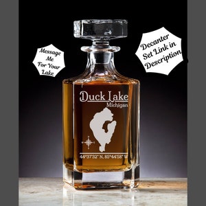 May include: A clear glass decanter with a square base and a stopper. The decanter is engraved with the words "Duck Lake, Michigan" and the coordinates "44°37'32" N, 85°44'58" W".