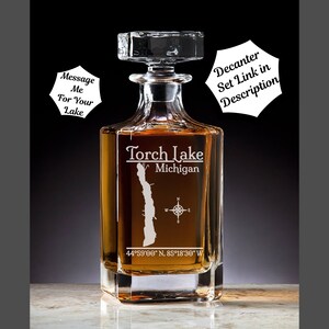 May include: A clear glass decanter with a square base and a stopper. The decanter is etched with an outline of Torch Lake, Michigan, and the coordinates 44°59'00" N, 85°18'30" W. The text "Torch Lake Michigan" is also etched on the decanter.