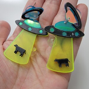 May include: A pair of neon green and black UFO earrings with a black cow silhouette on each earring.