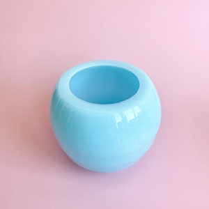 以下が含まれることがあります： 滑らかで丸みを帯びた形をした、薄い青色のセラミック製のプランター。プランターは空で、植物を入れる準備ができています。