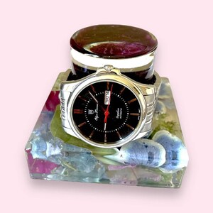 May include: A silver watch with a black face and a date window that reads "THU 14". The watch is sitting on a square resin block with embedded flowers.